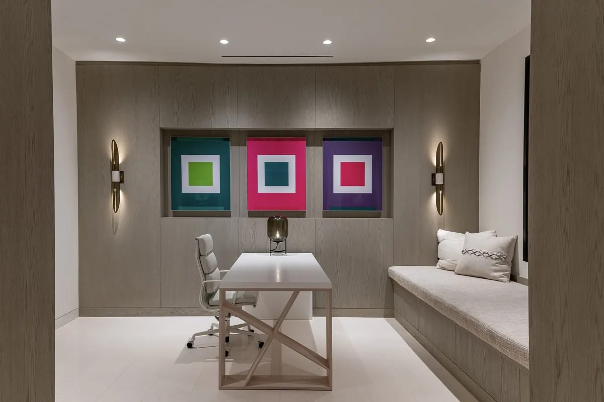 Office with light wood desk, white chair, built-in bench, and colorful framed wall art