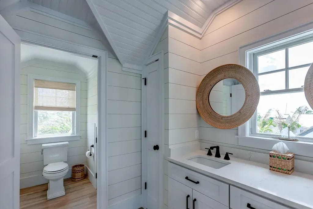Bathroom with toilet, sink, black faucets, round mirror, white shiplap walls, and light wood flooring.