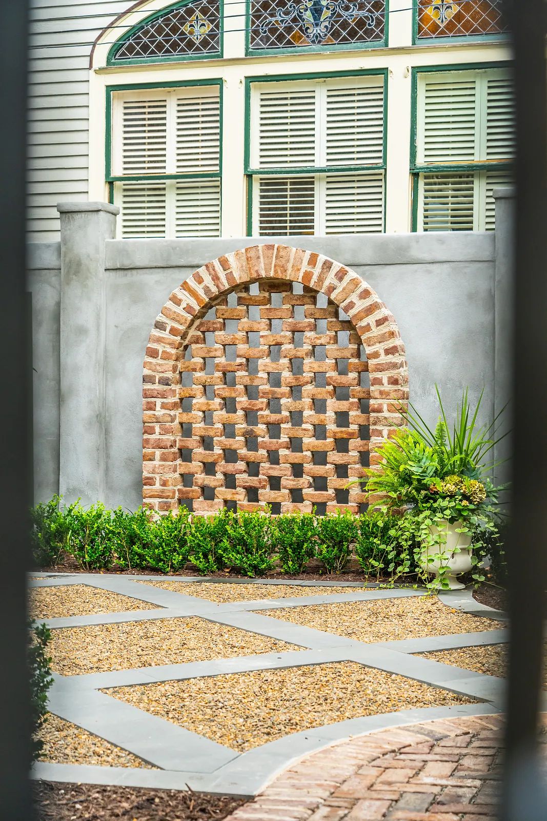 Garden area with brick lattice wall, gravel walkway, and manicured shrubbery around a potted plant.