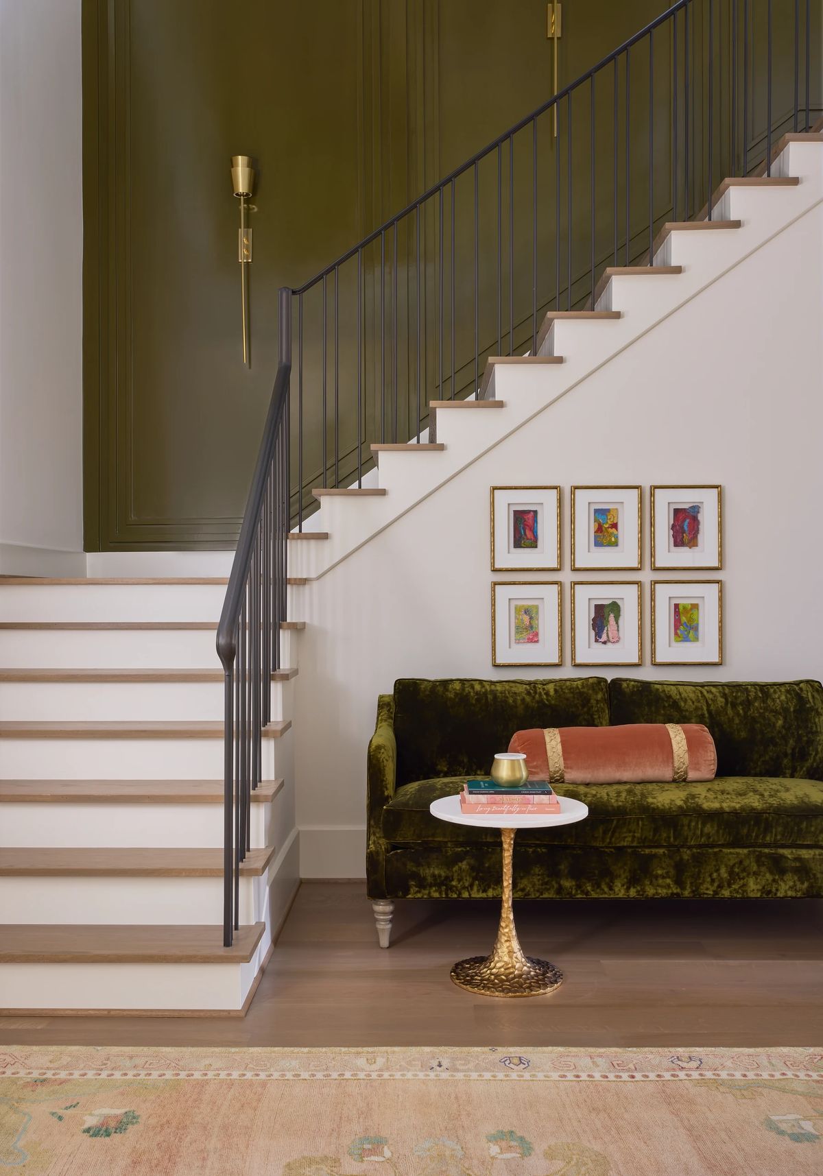 Living room with green velvet sofa, white side table, gold accent table, and staircase with artwork on the wall.