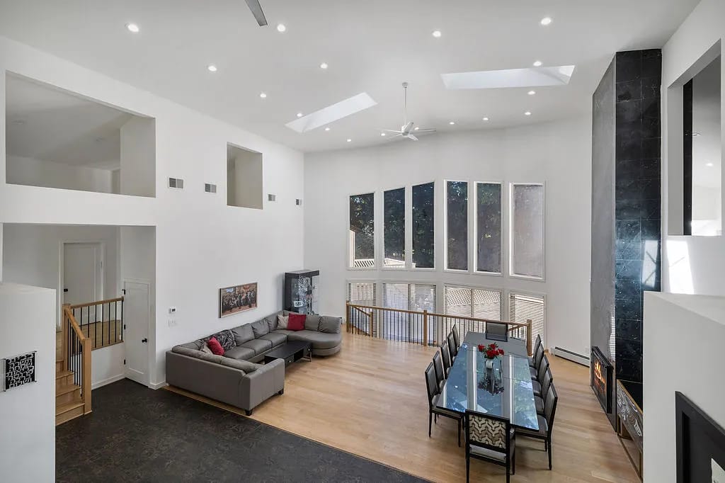 Spacious living room with gray sectional sofa, black dining table, large windows, fireplace, and high ceiling with skylights