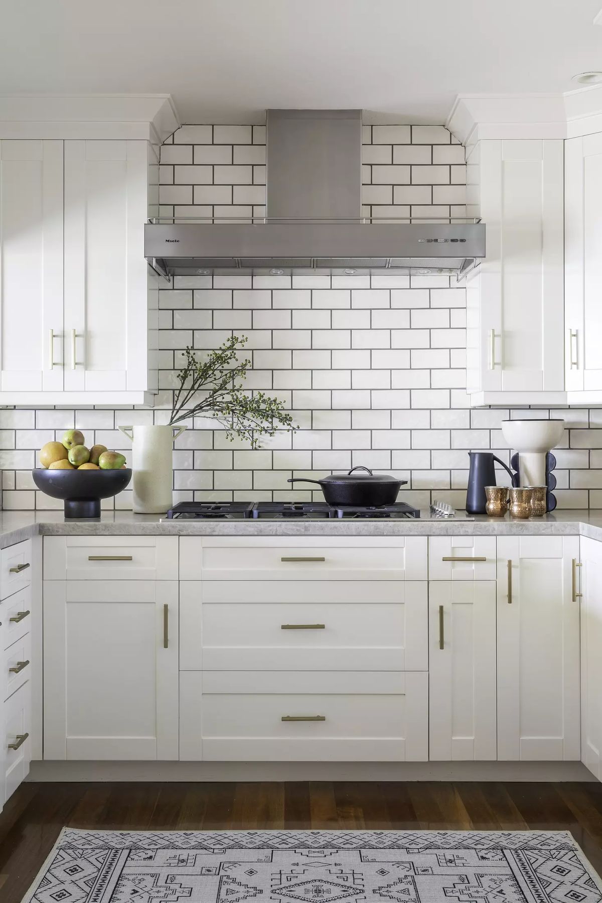 Kitchen with white cabinets, gold hardware, stainless steel range hood, fruit bowl, and patterned rug on wooden floor.