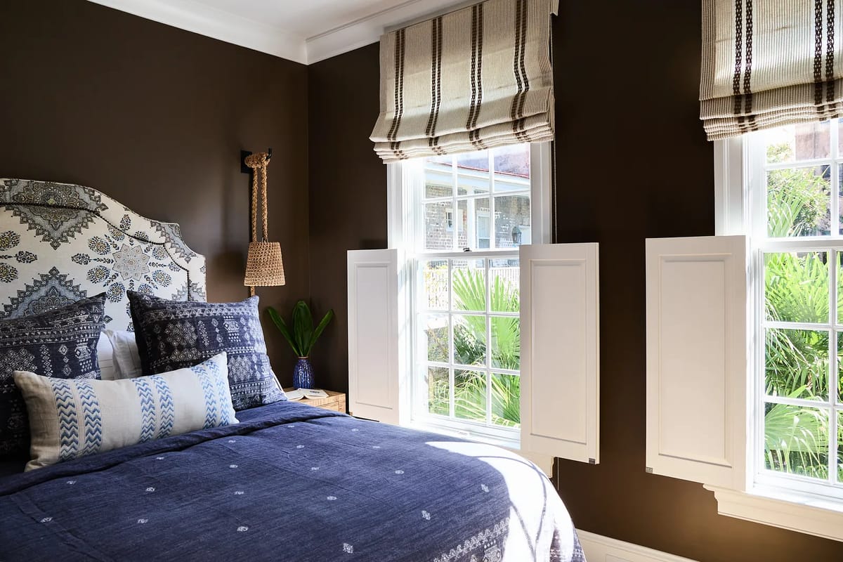 Bedroom with patterned headboard, blue bed linens, windows with shutters, and nightstand with plant.