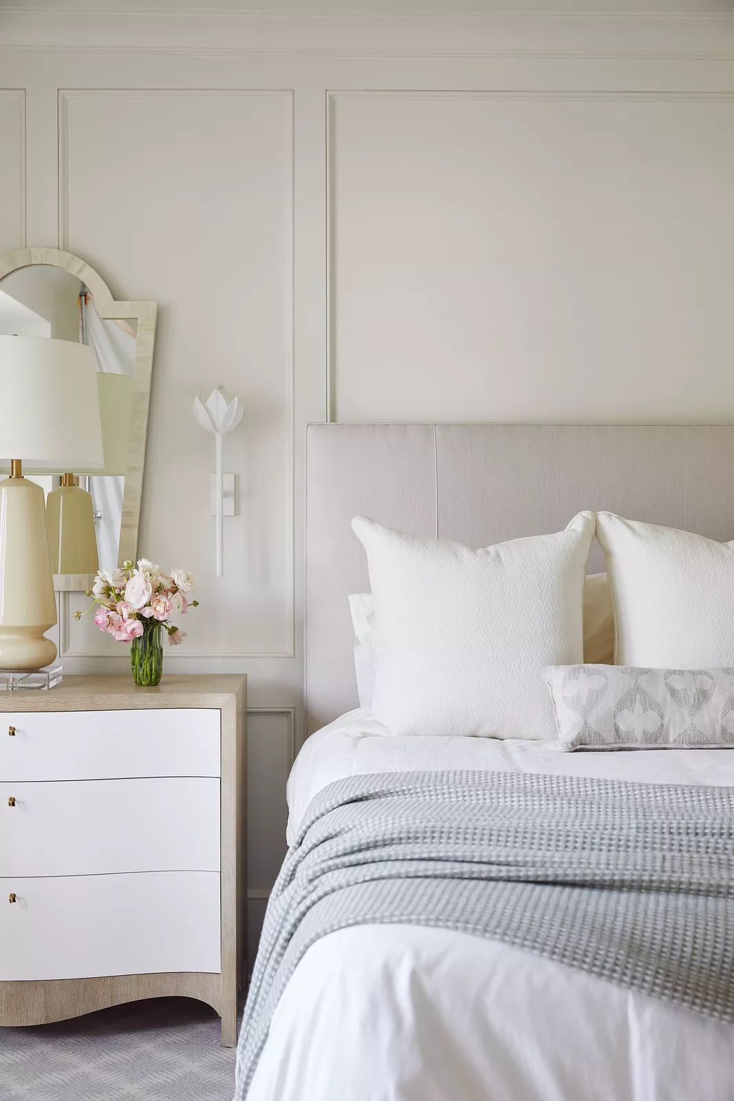 Bedroom with white bedding, beige headboard, nightstand, vase of flowers, and wall paneling.
