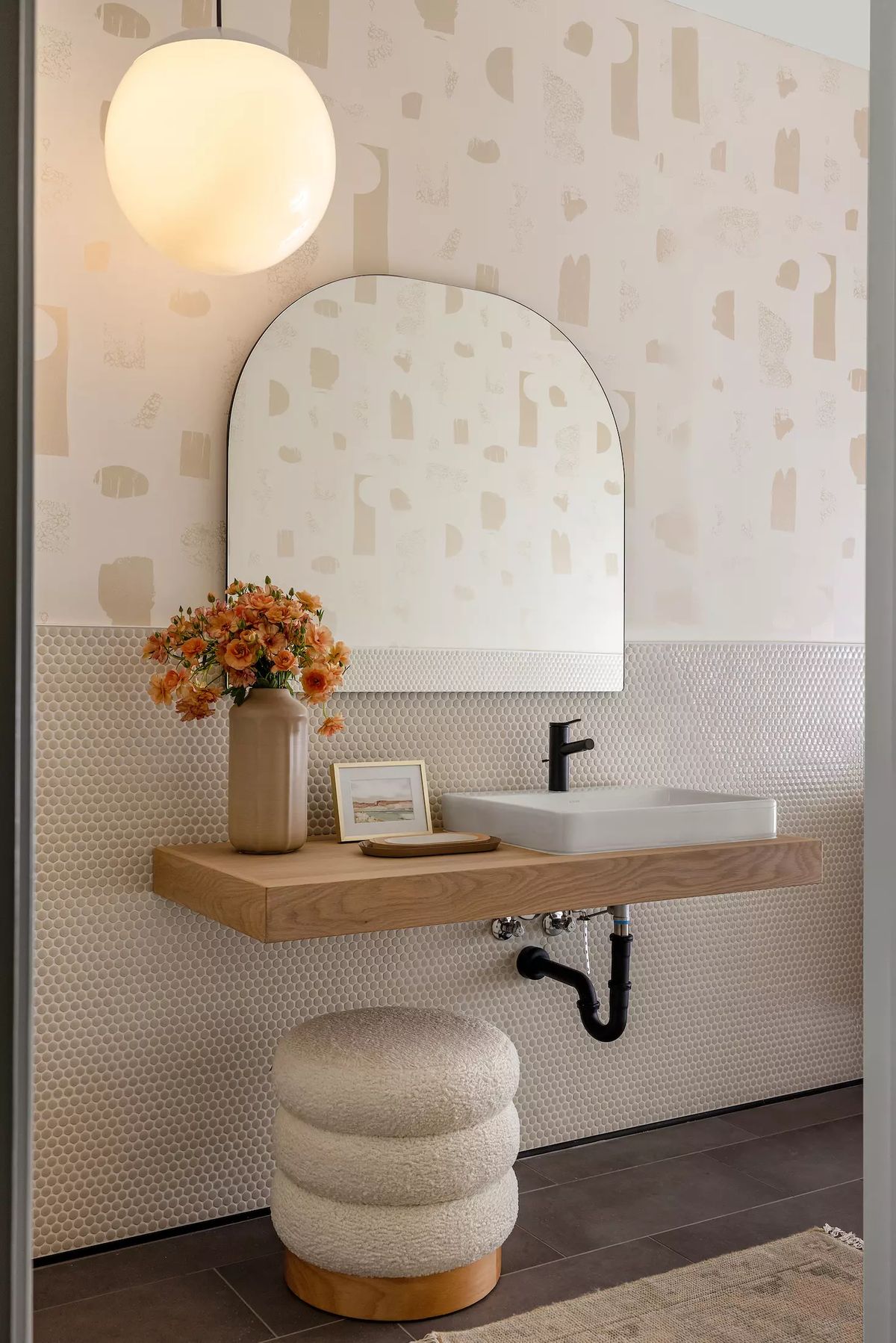 Powder room with wall-mounted sink, round mirror, wooden shelf, patterned wallpaper, and textured stool
