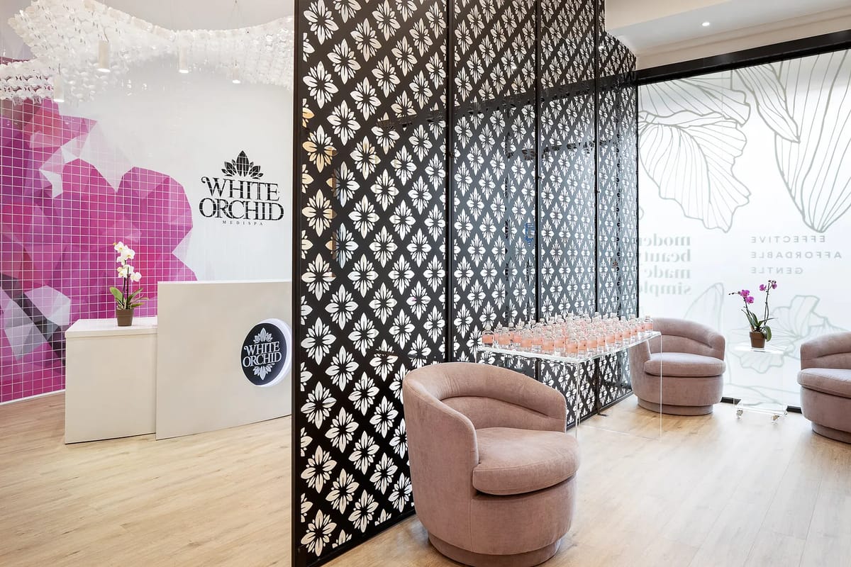 Reception area with white desk, pink tiled wall, patterned screen, rounded chairs, and drinks on glass table.
