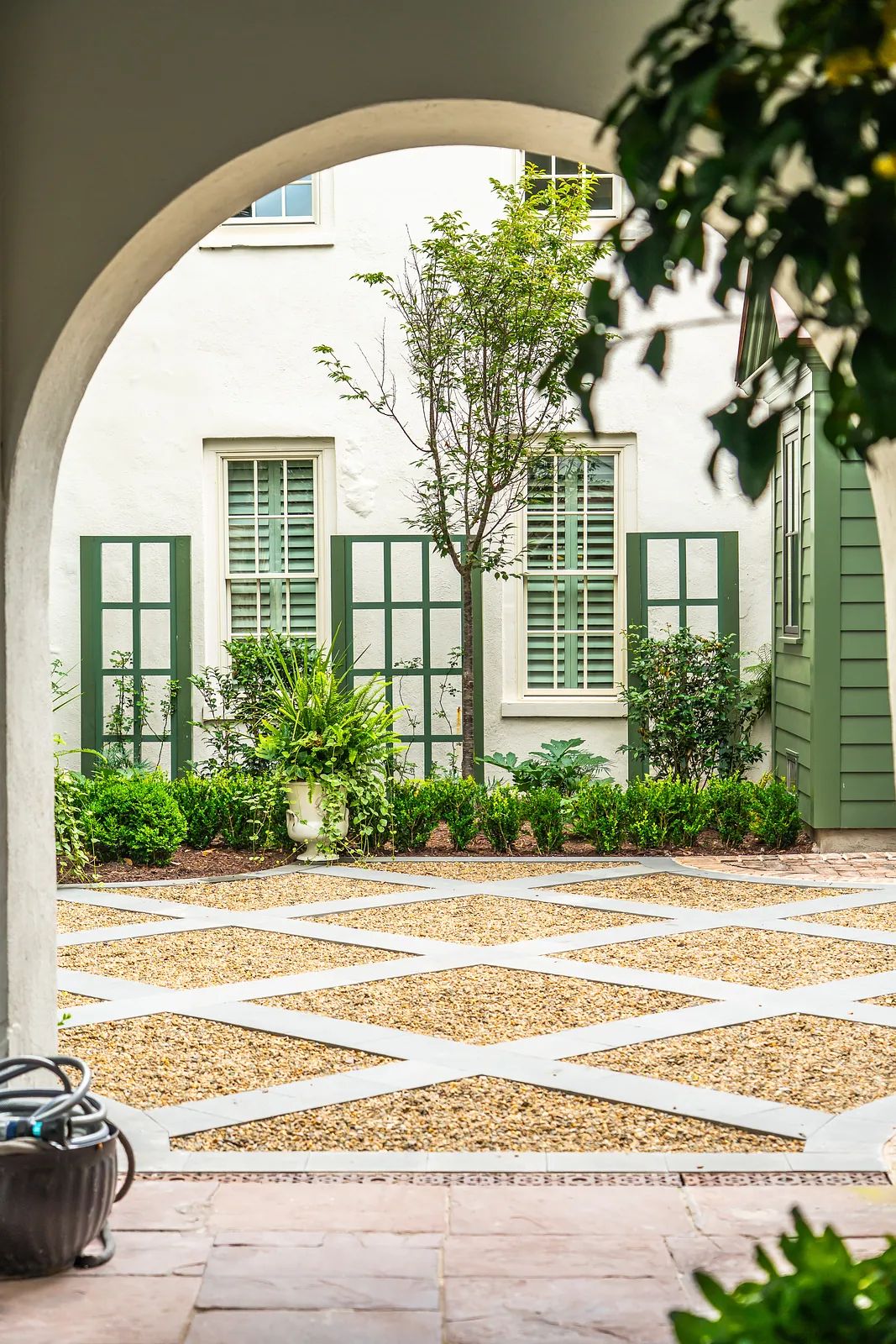 Garden area with gravel pathway, greenery, potted plants, and white walls with green shutters.