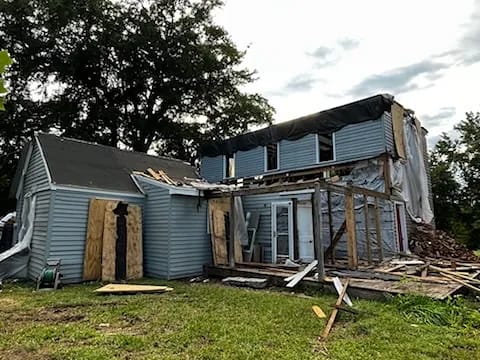 Partially demolished house with blue siding, exposed framing, tarp-covered roof, and debris in the yard.