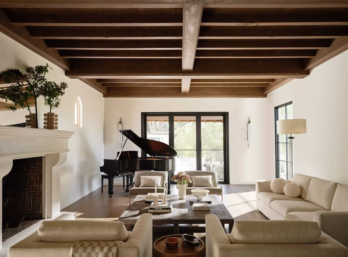 Living room with grand piano, beige sofas, coffee table, fireplace, wooden beams, and large windows