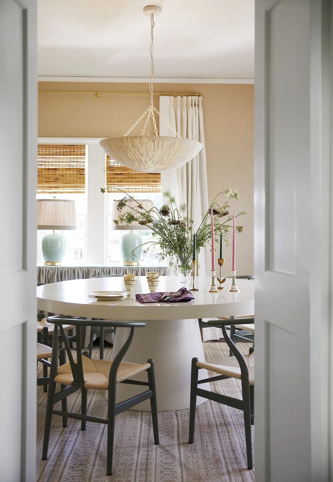 Dining room with round white table, black chairs, pendant light, vases, candles, and neutral walls.
