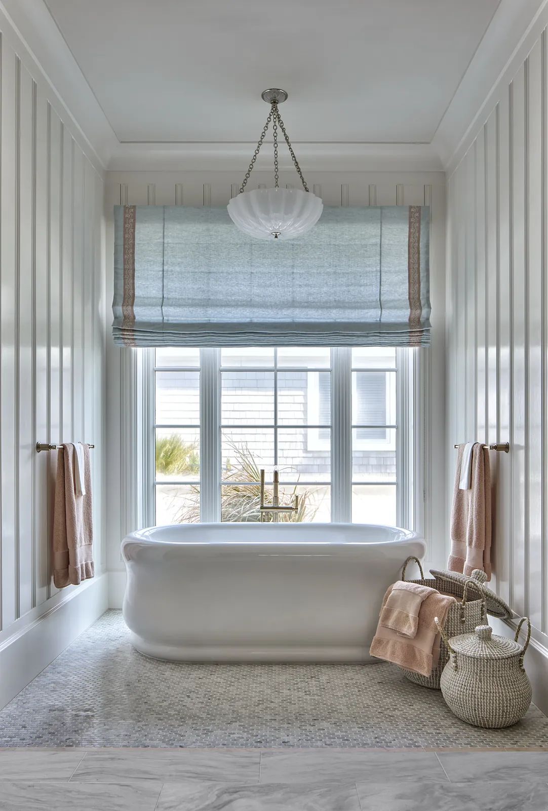 Bathroom with freestanding white bathtub, stone floor, blue window treatment, and pastel towels.