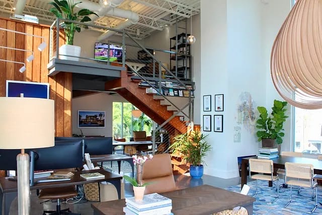 Office space with wooden desk, computer, plants, staircase, shelving unit, and natural light from large windows
