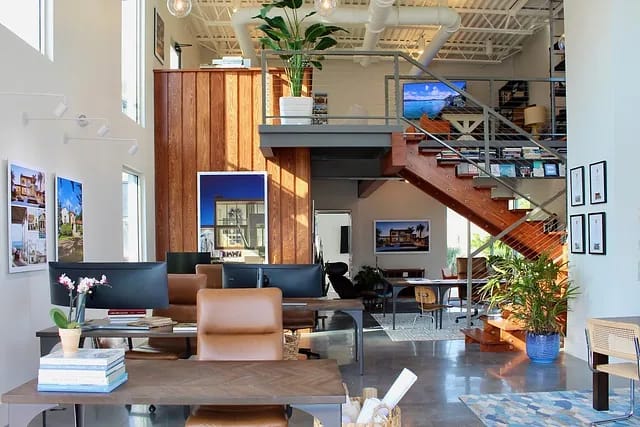 Office space with desks, chairs, staircase, large windows, wooden accents, and indoor plants.
