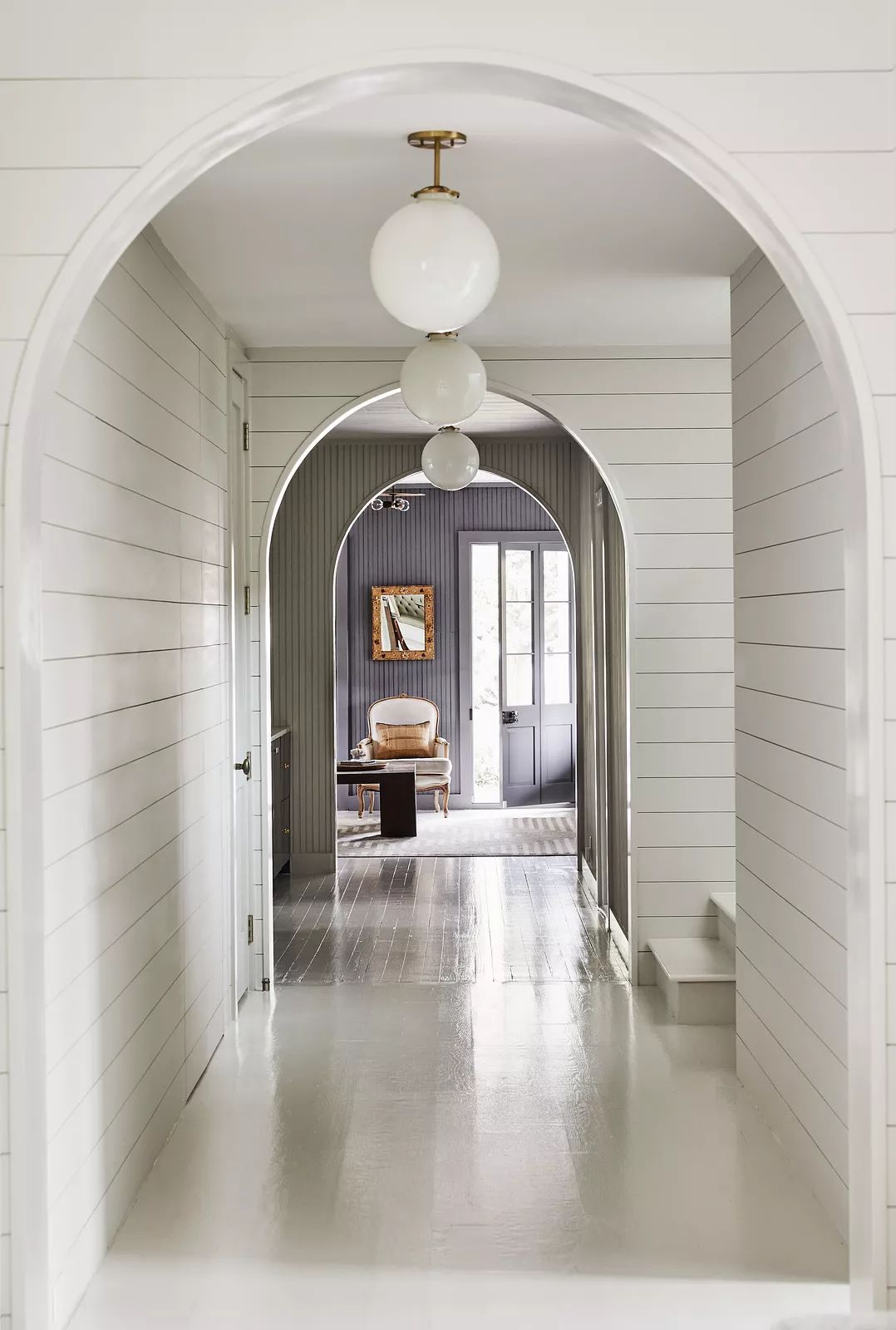 Hallway with white shiplap walls, glossy floor, arched doorways, and three spherical ceiling fixtures.