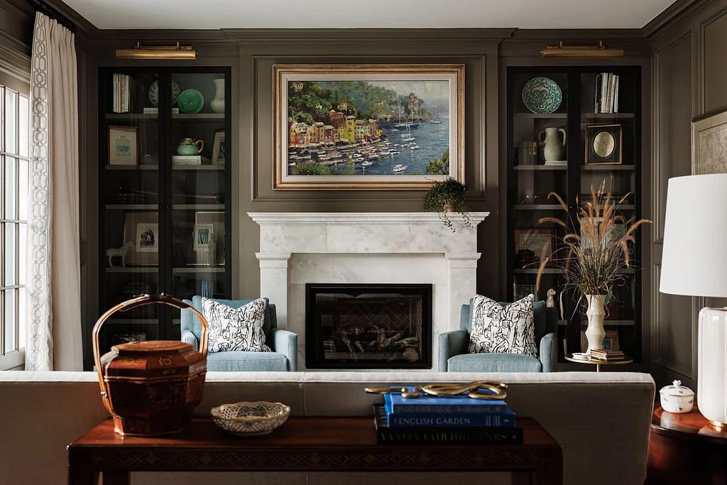 Living room with blue armchairs, marble fireplace, dark cabinetry, coastal painting, and console table with books.