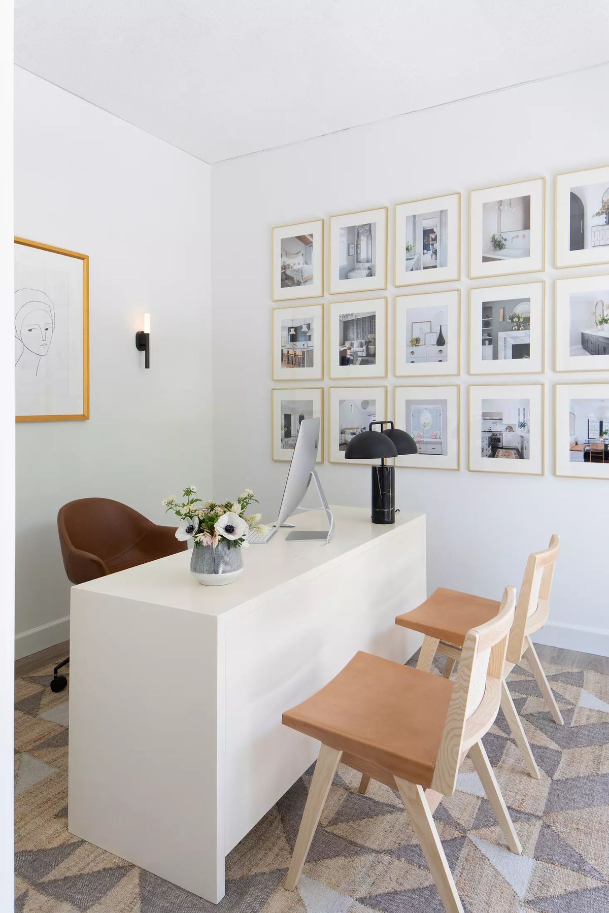 Office with white desk, brown chair, two light wood chairs, black lamp, flower arrangement, and gallery wall.
