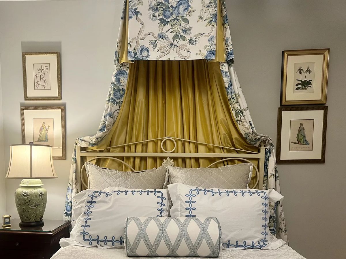 Bedroom with canopy bed, cream linens, decorative pillows, framed artwork, and bedside lamp.
