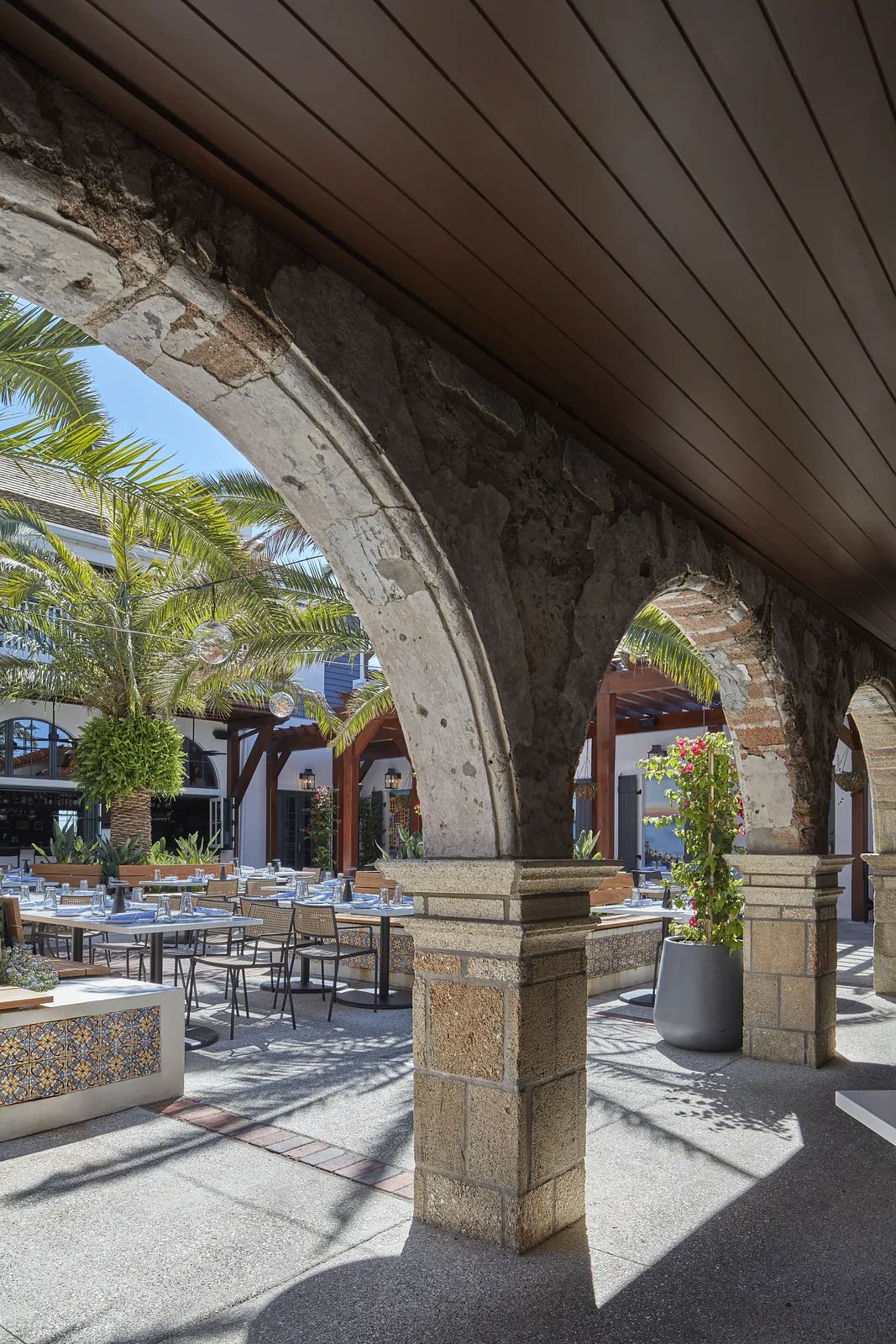Outdoor patio with stone archways, wooden beams, dining tables, chairs, and potted plants.