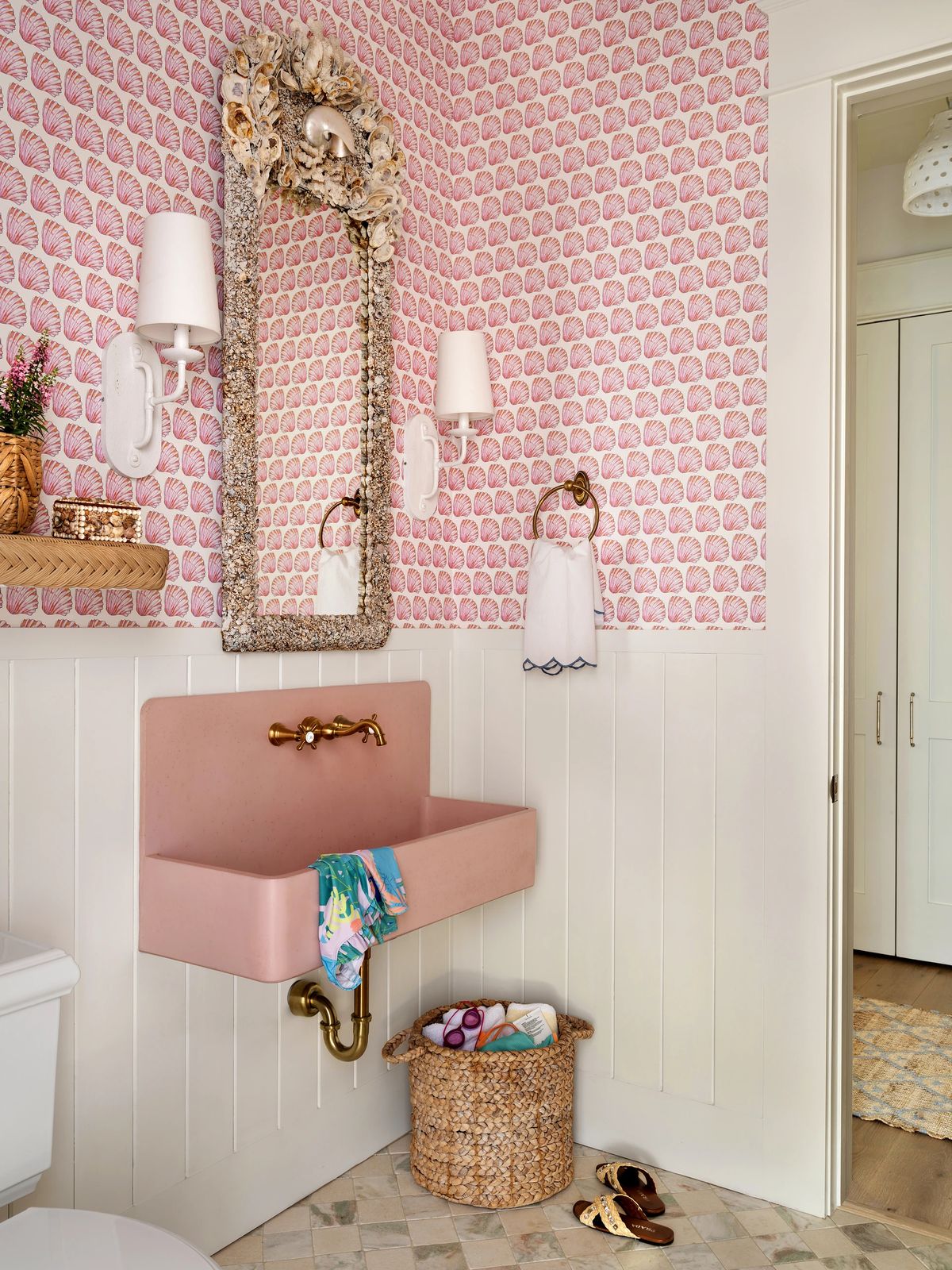 Powder room with pink sink, floral wallpaper, white wainscoting, mirror, and woven basket on tiled floor.