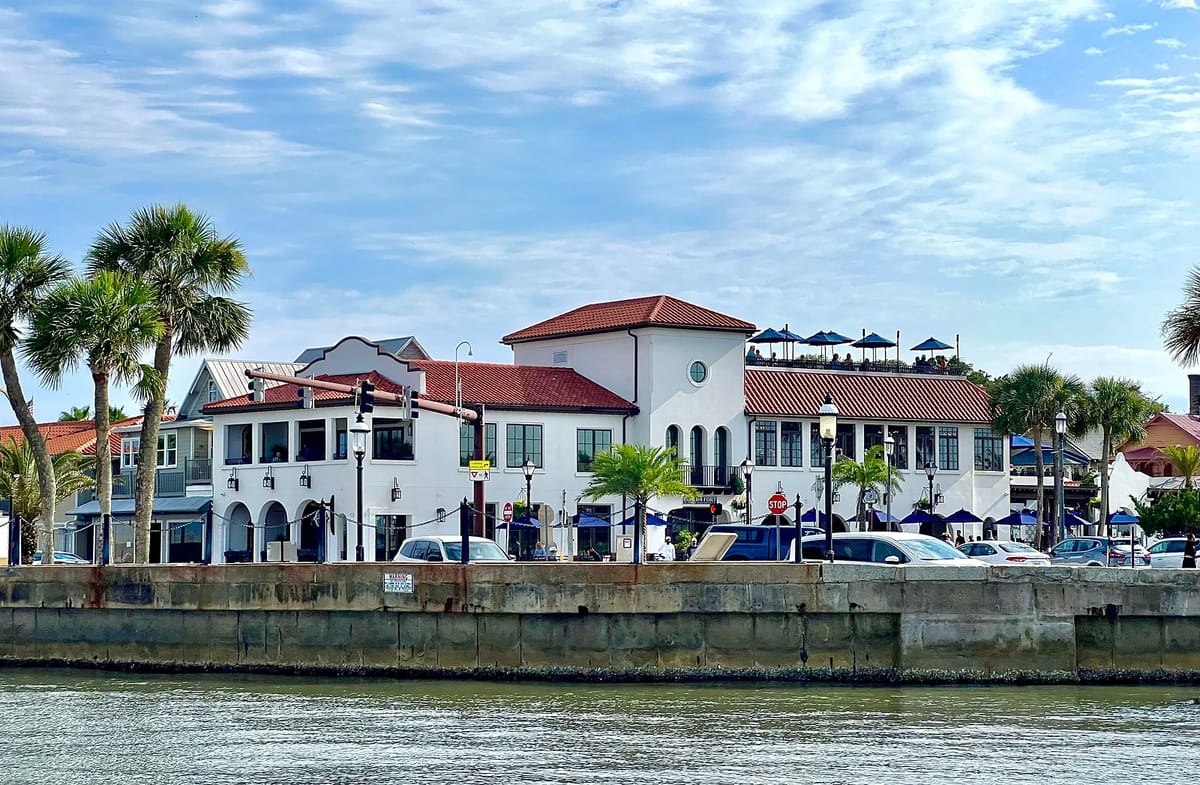 Waterfront view of a two-story building with white facade, red tile roof, balconies, and palm trees.