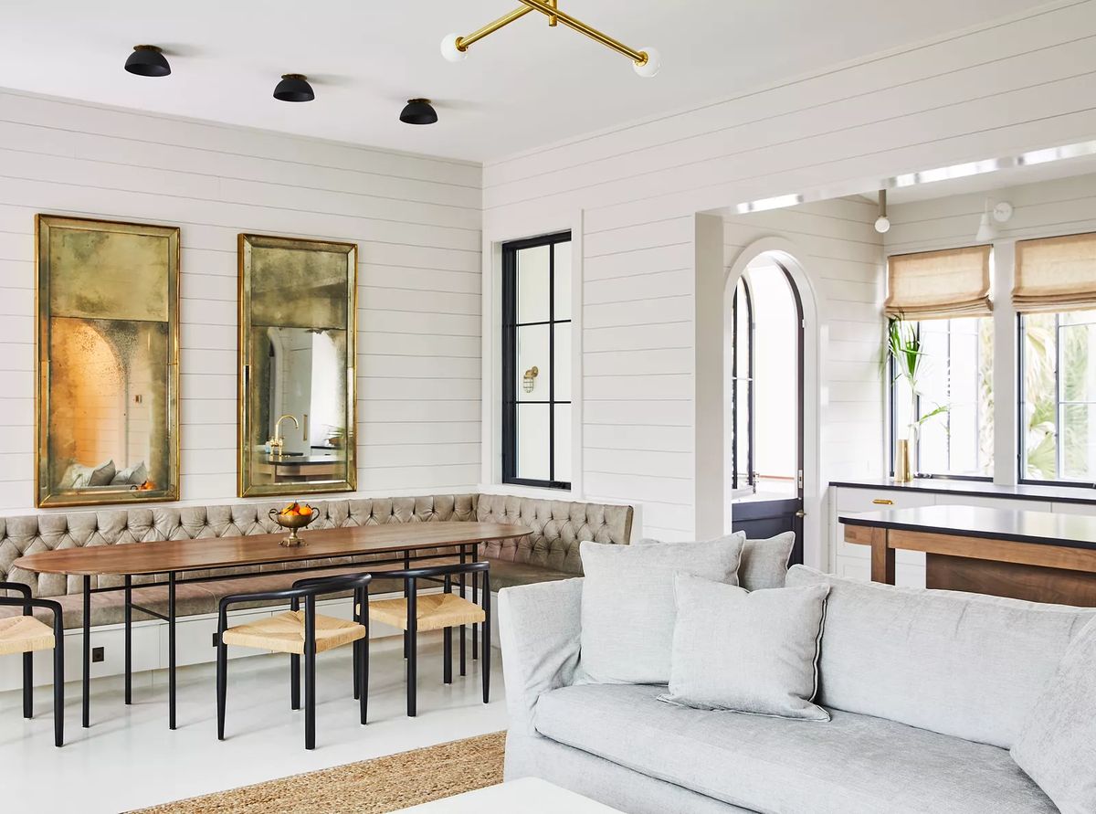 Dining room with wooden table, beige banquette, mirrors, and shiplap walls providing natural light.