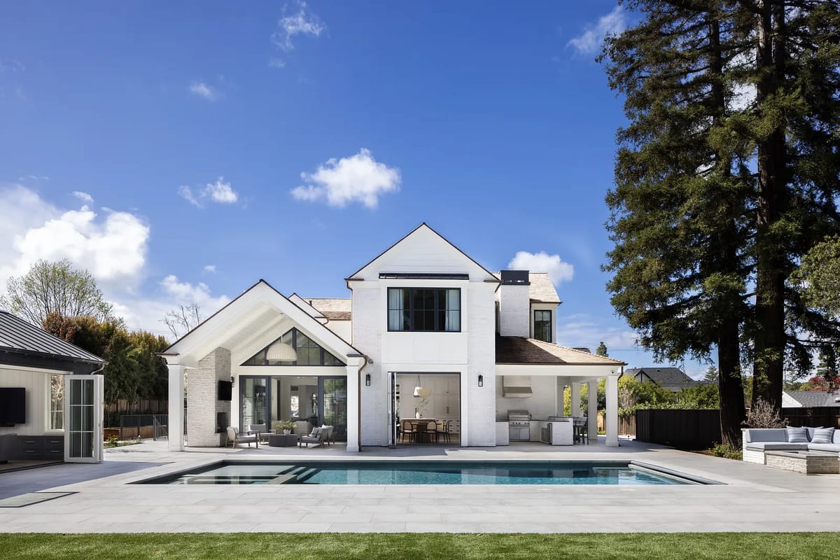 Rear exterior view of a house with a swimming pool, patio, large windows, and tall trees.