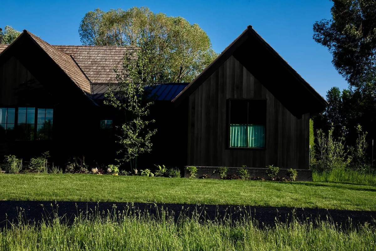Front exterior of house with dark wood siding, large windows, and green curtains in the landscape.