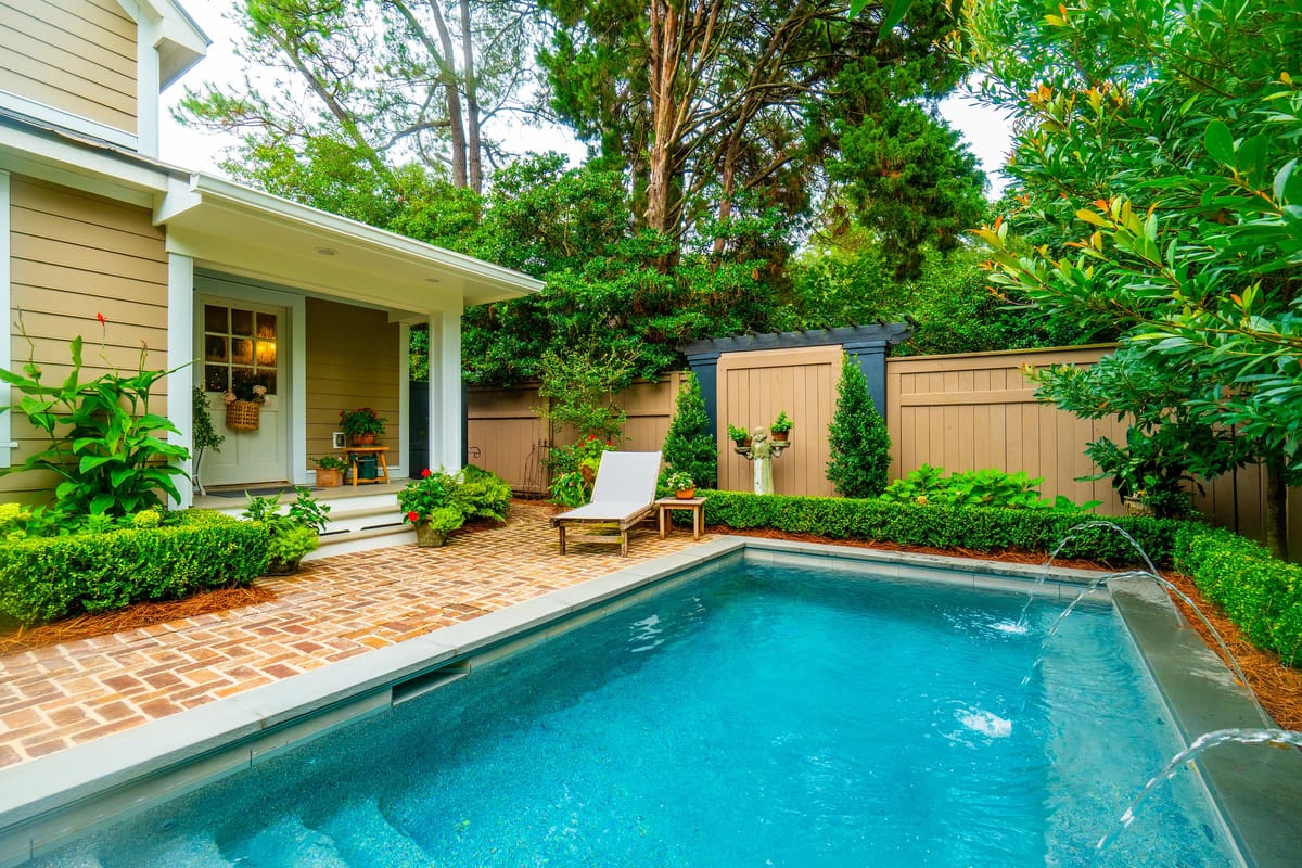 Patio with swimming pool, lounge chair, wooden tables, and potted plants surrounded by greenery.