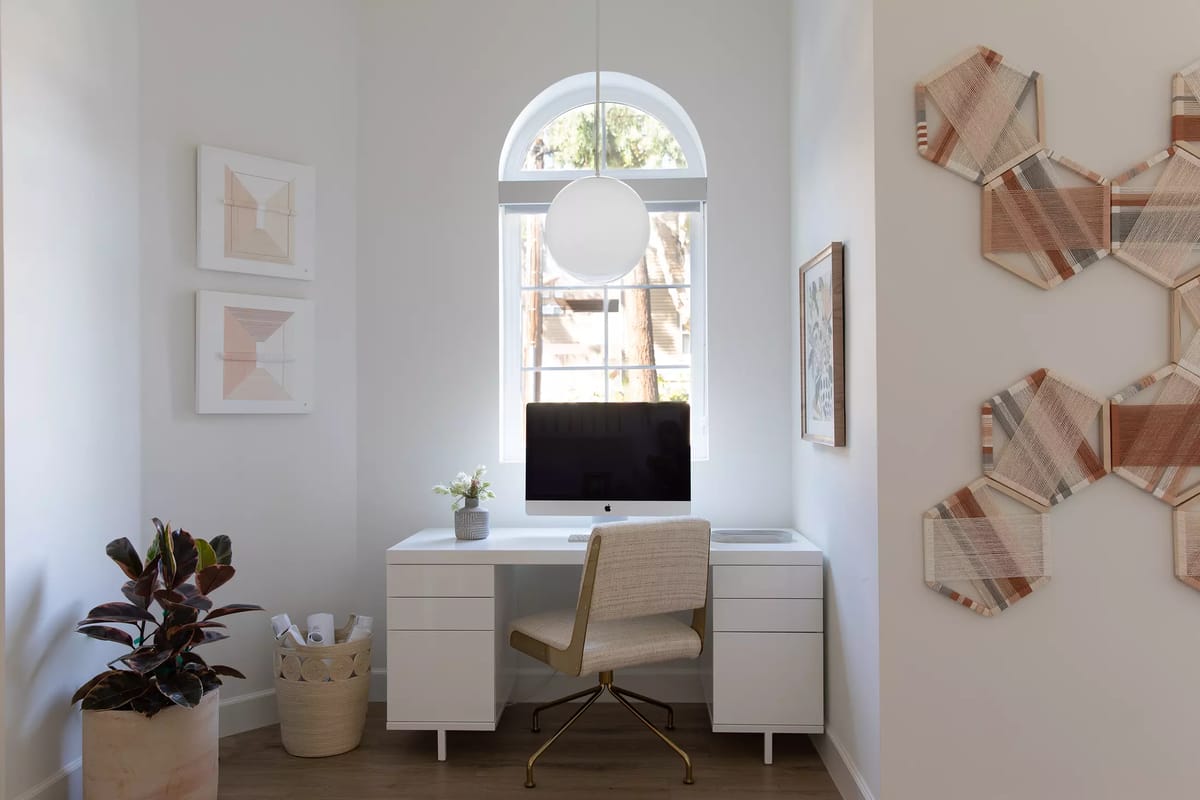 Office with white desk, beige chair, computer, wall art, potted plant, and arched window.