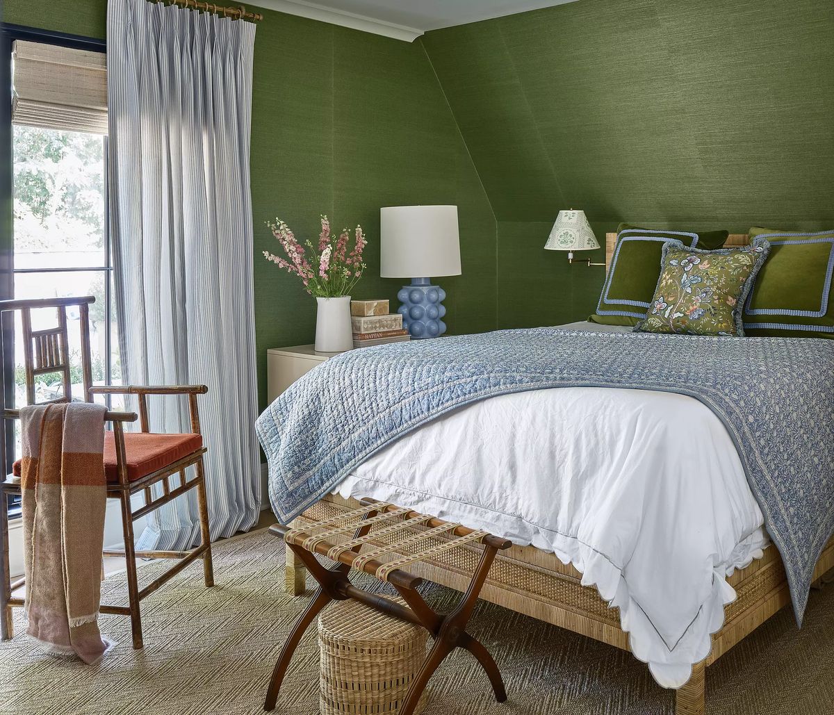 Bedroom with bed, nightstand, blue lamp, wicker stool, chair, and curtain over window with natural light.