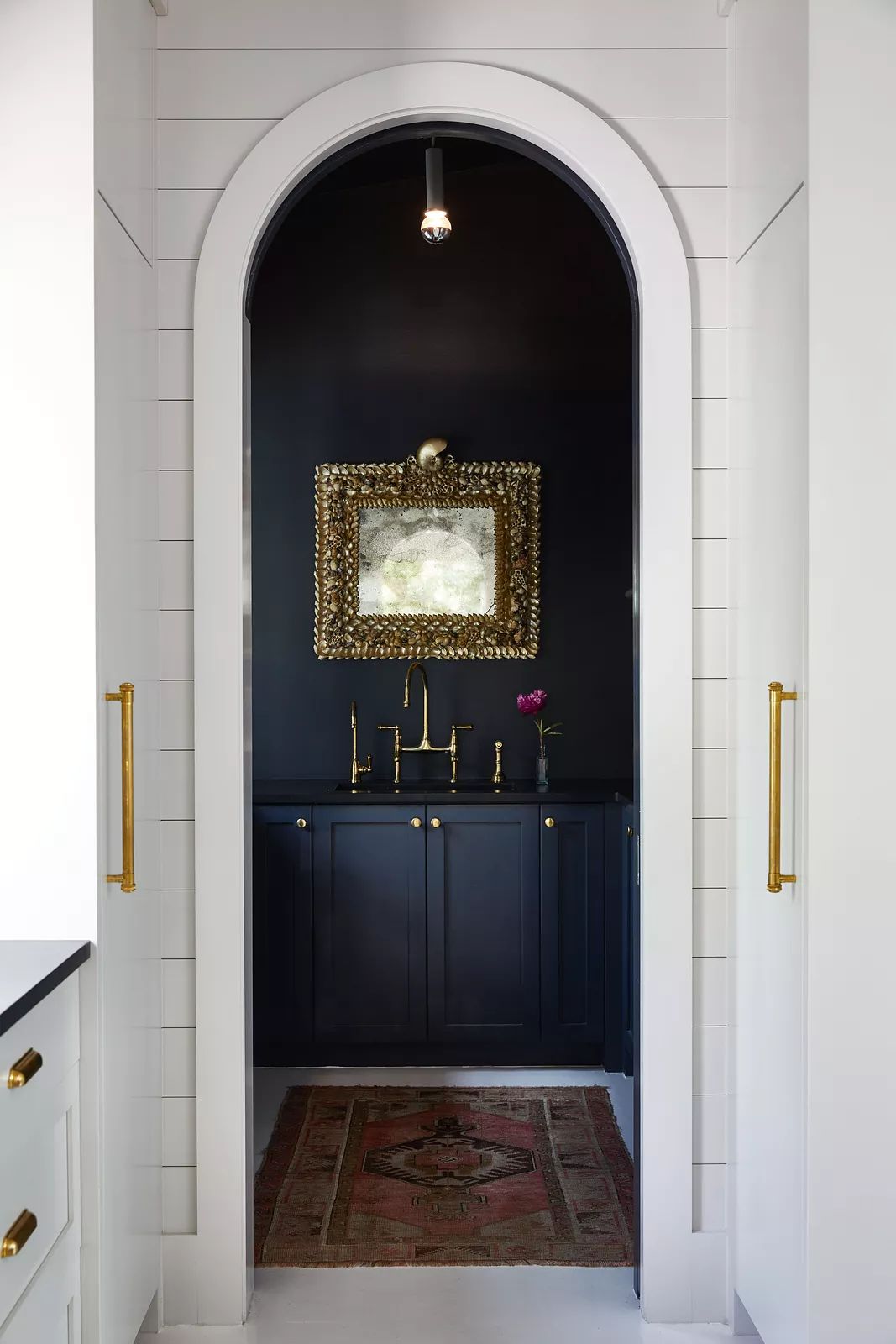 Powder room with dark wall, gold-framed mirror, black cabinetry, brass fixtures, and vintage rug.