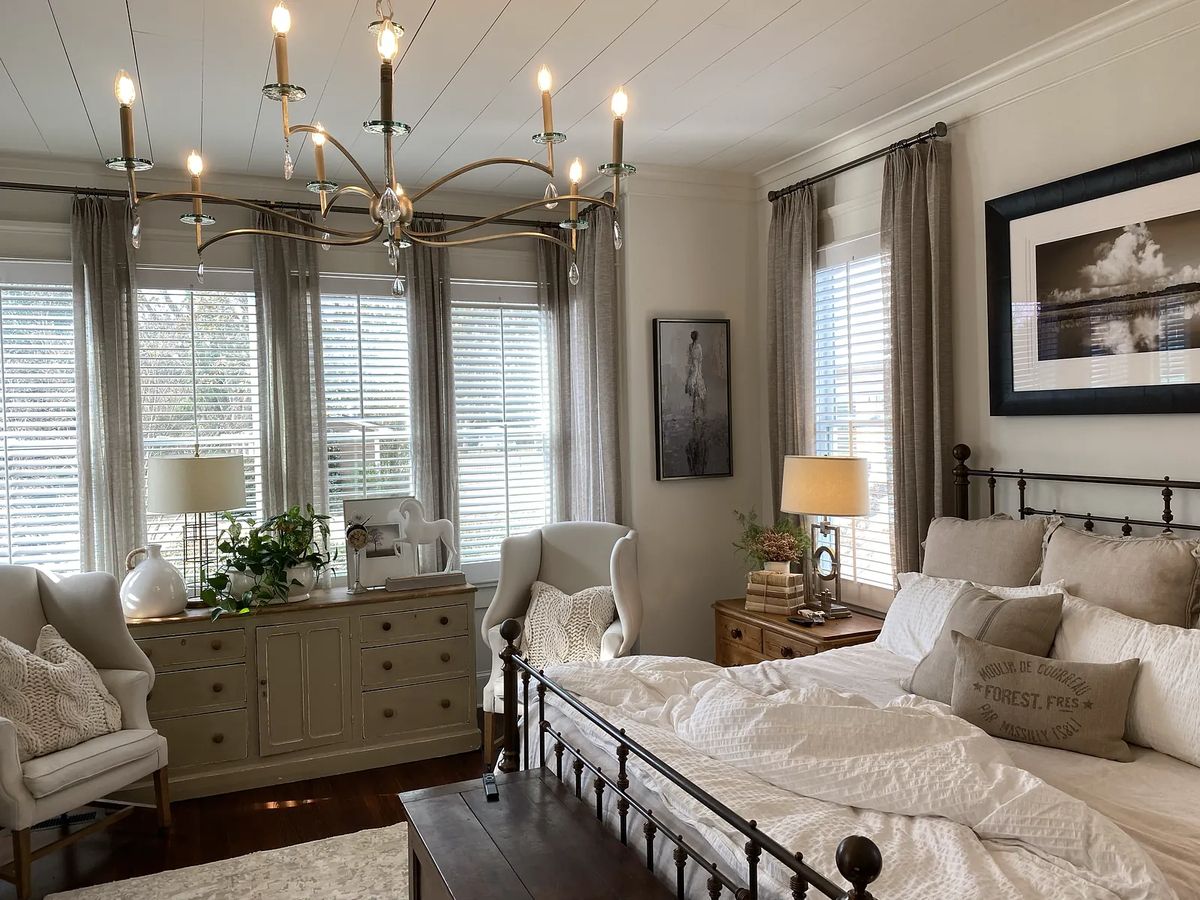 Bedroom with metal bed, armchairs, dresser, chandelier, large windows, and decorative elements.