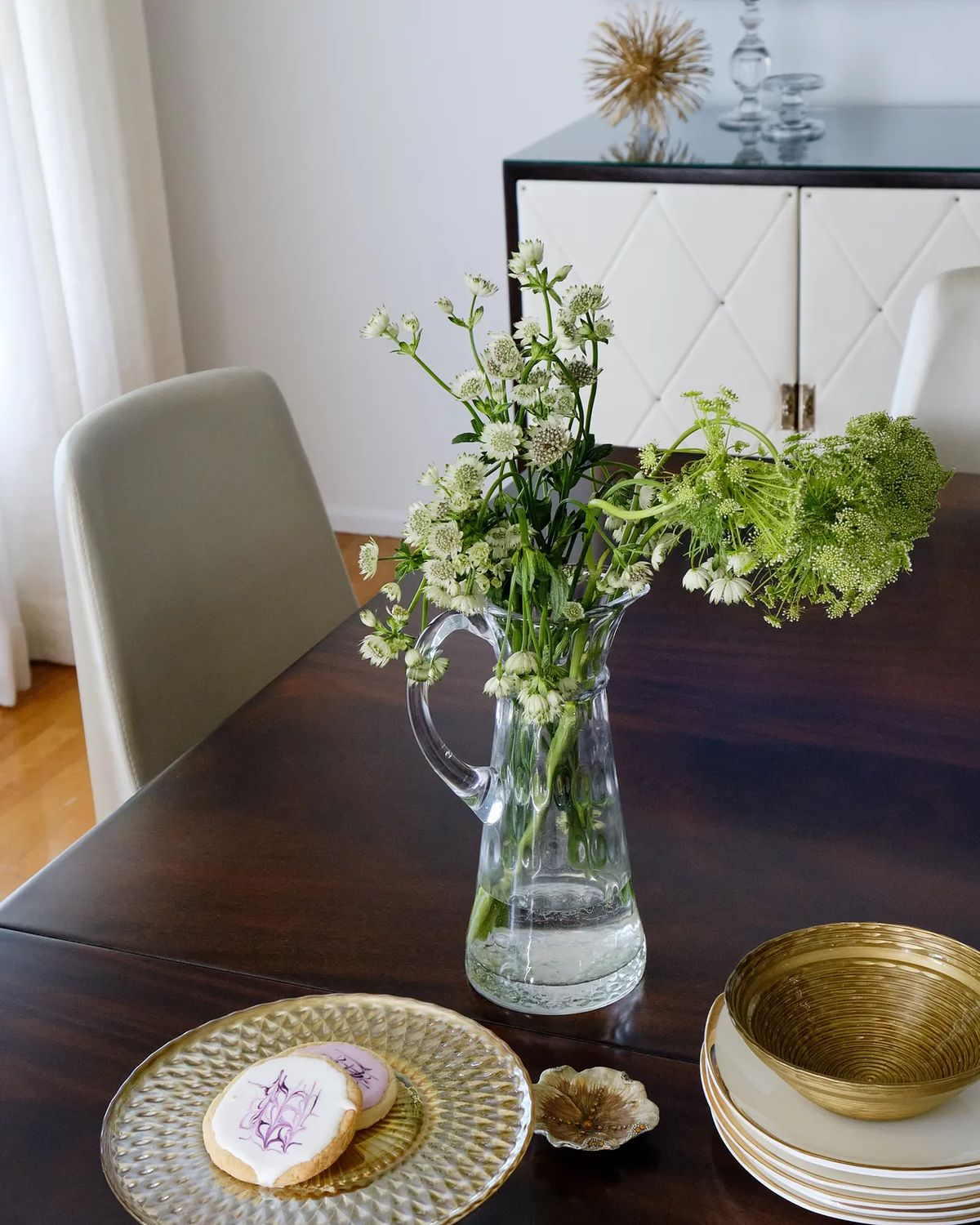 Dining room with wooden table, flowers, decorative plates, and a quilted cabinet in the background.