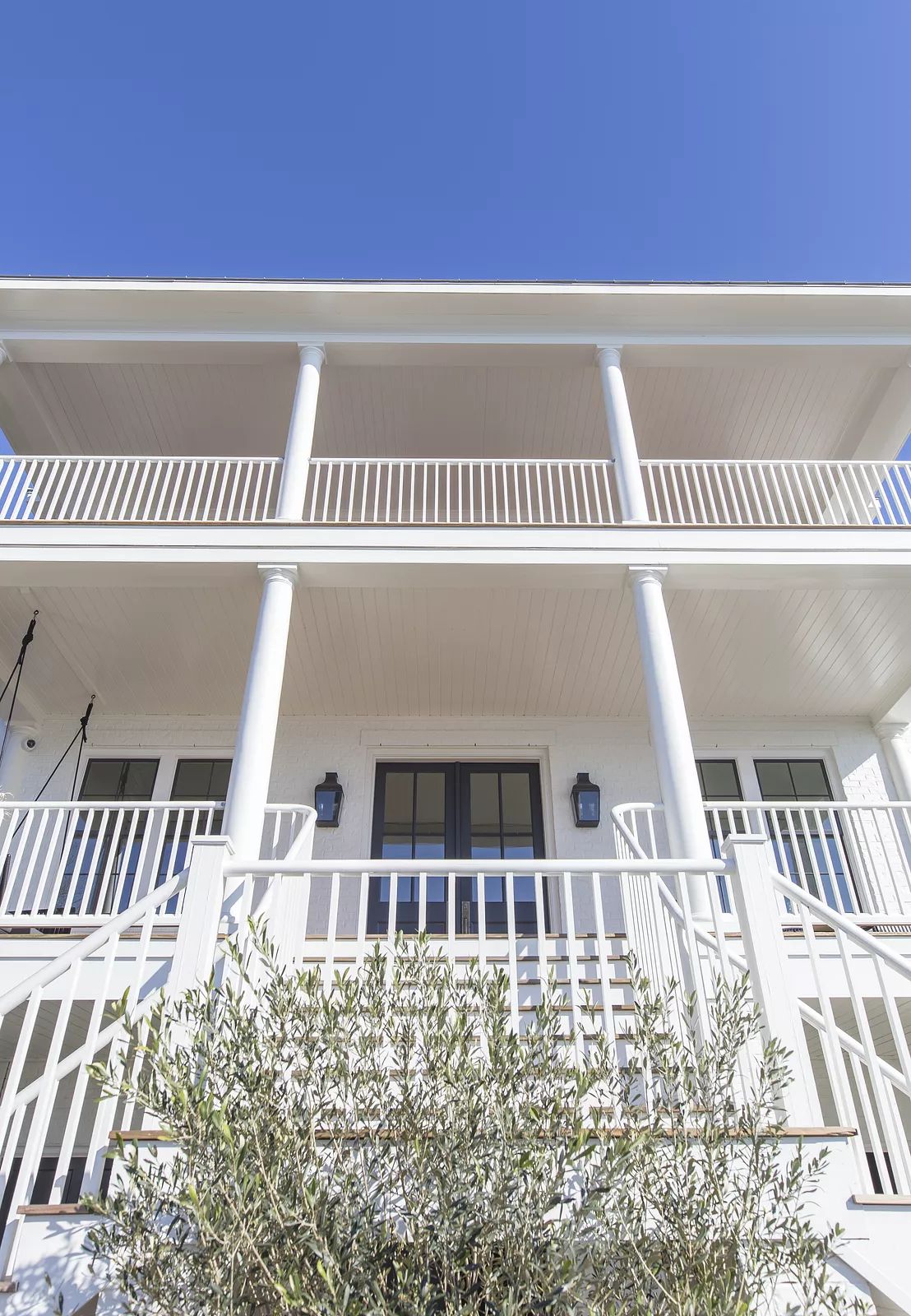 Exterior front view with white pillars, balcony, lanterns, large windows, and green bushes.