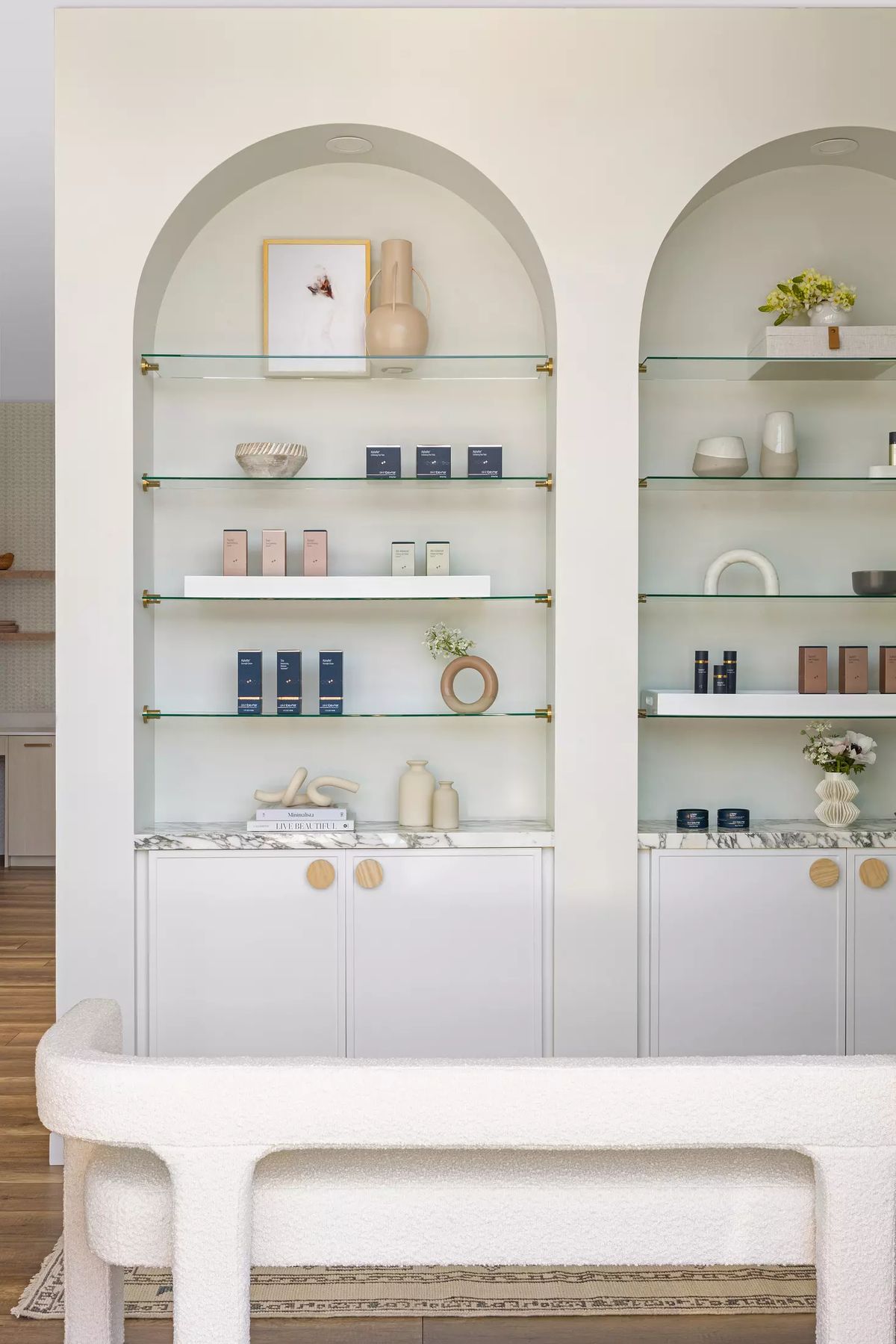 Display area with arched glass shelves, skincare products, cabinet below, and a textured bench.