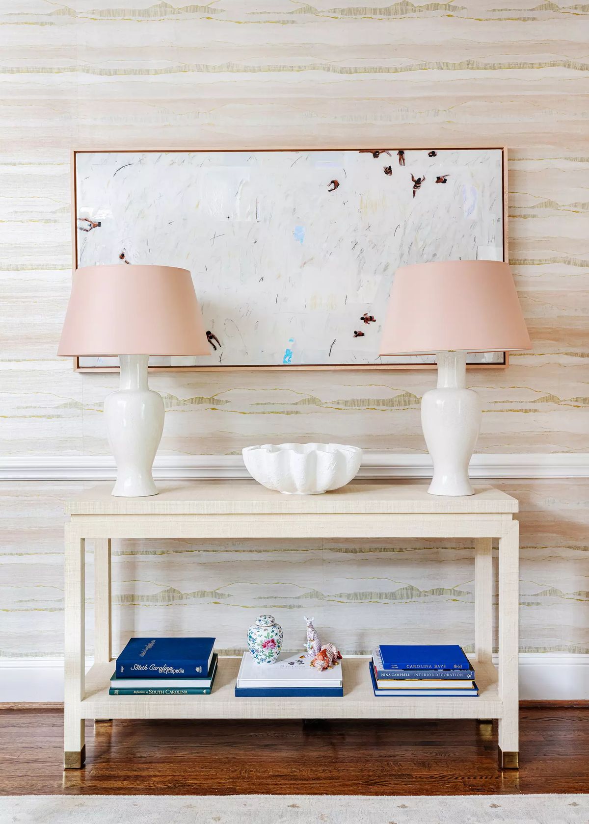 Hallway with console table, pink lamps, decorative bowl, stacked books, and large framed artwork on striped wallpaper