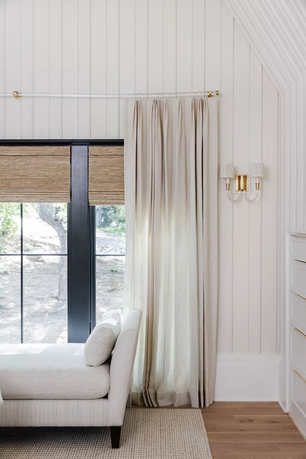 Seating area with chaise lounge, sheer curtains, large window, natural fiber rug, and wall-mounted sconces.