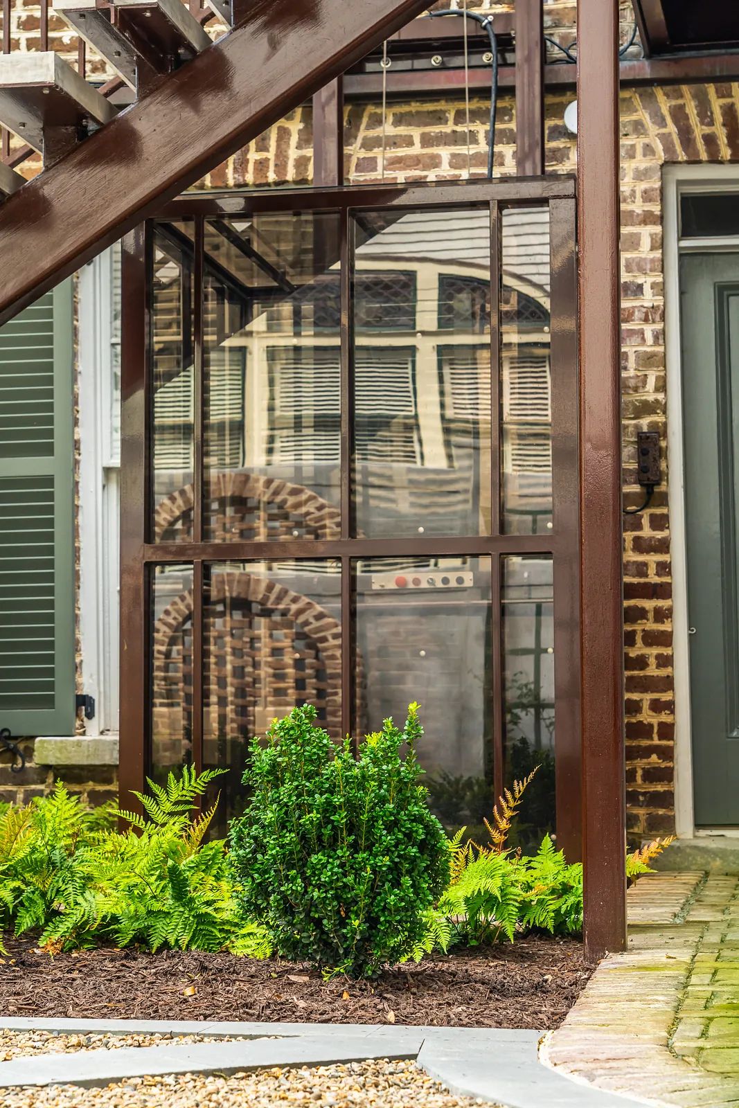 Garden area with glass enclosure, green shrub, ferns, gravel path, and wooden staircase.