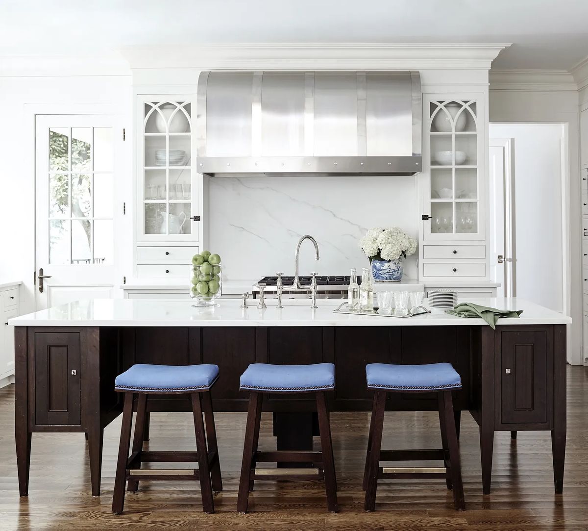 Kitchen with central island, blue stools, stainless steel range hood, marble backsplash, and wooden cabinetry.