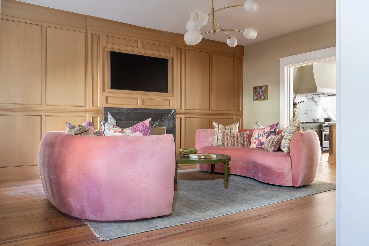 Living room with pink curved sofas, green coffee table, gray rug, and light wood-paneled wall with TV.