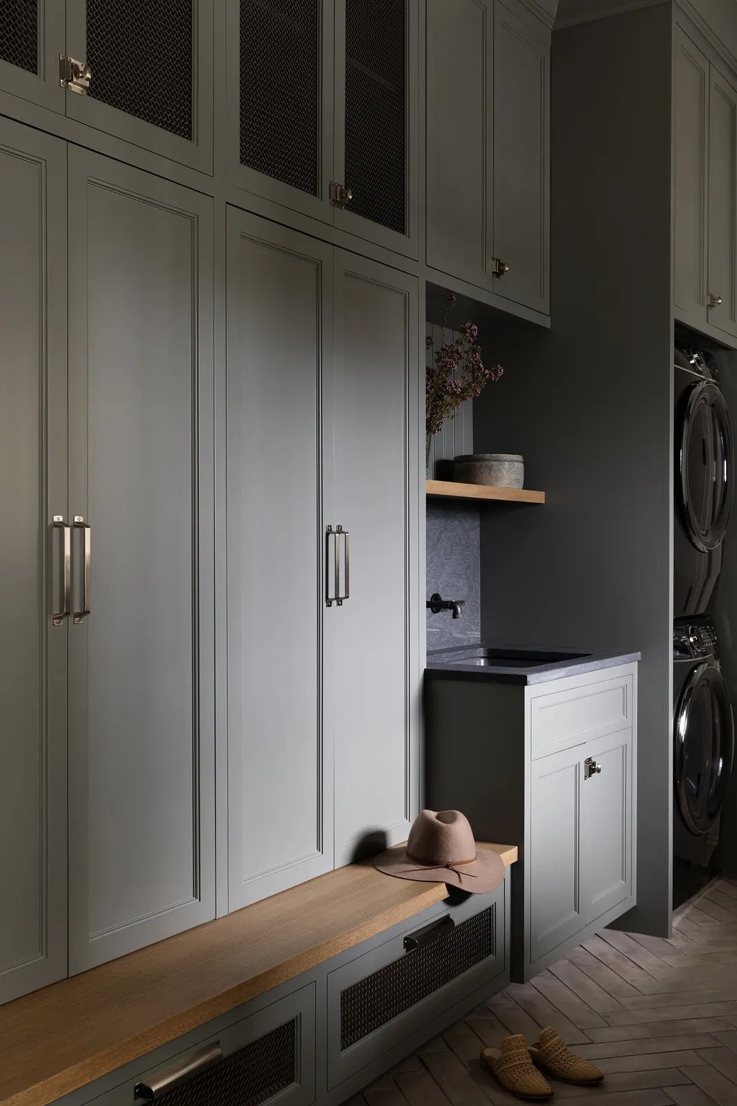 Laundry room with gray cabinetry, black countertop, wooden bench, washing machine, dryer, and herringbone flooring.