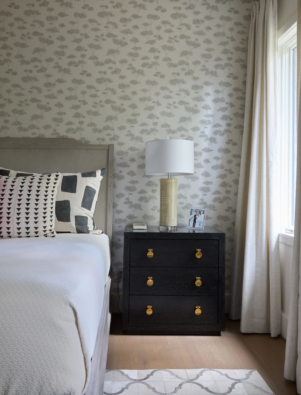 Bedroom with gray upholstered bed, black nightstand, table lamp, patterned wallpaper, and light curtains.