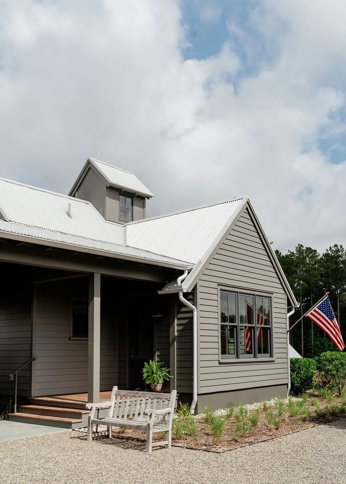 Exterior front of a house with gray wood facade, metal roof, front porch, bench, and American flag.