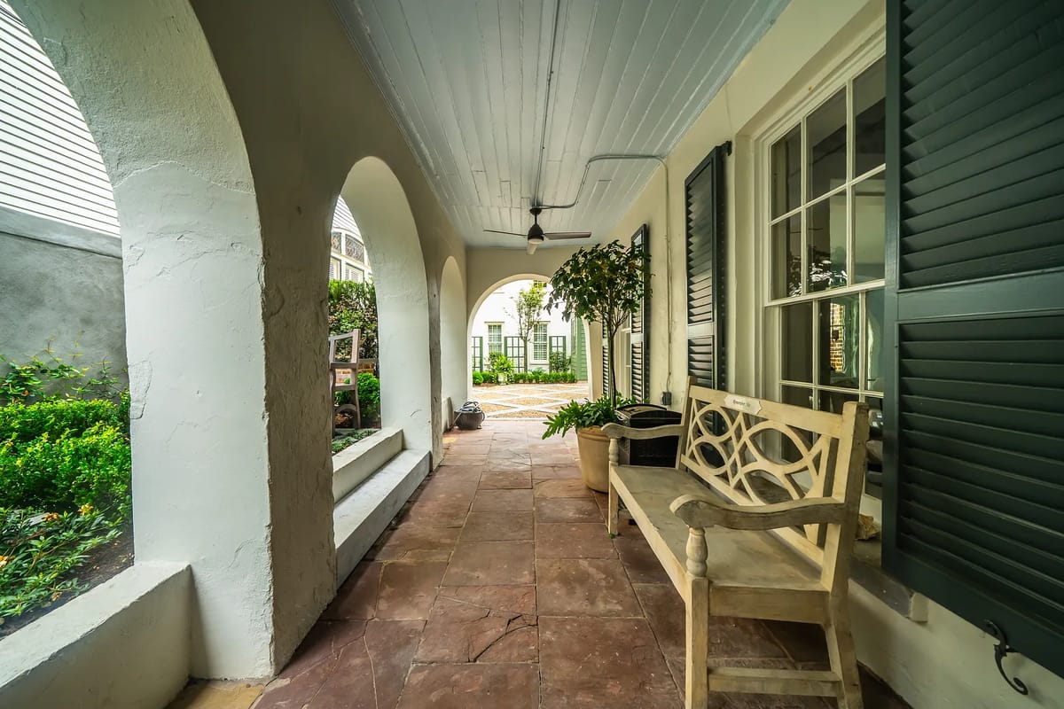 Exterior front space with stone patio, wooden bench, arched columns, and greenery.
