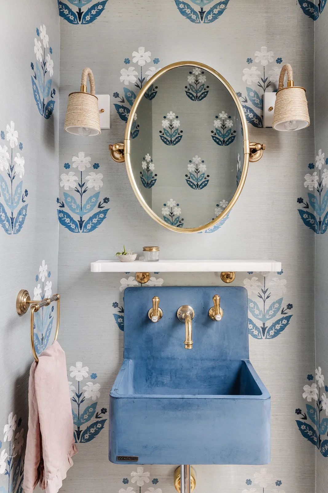 Powder room with blue sink, marble shelf, oval mirror, floral wallpaper, and brass fixtures.