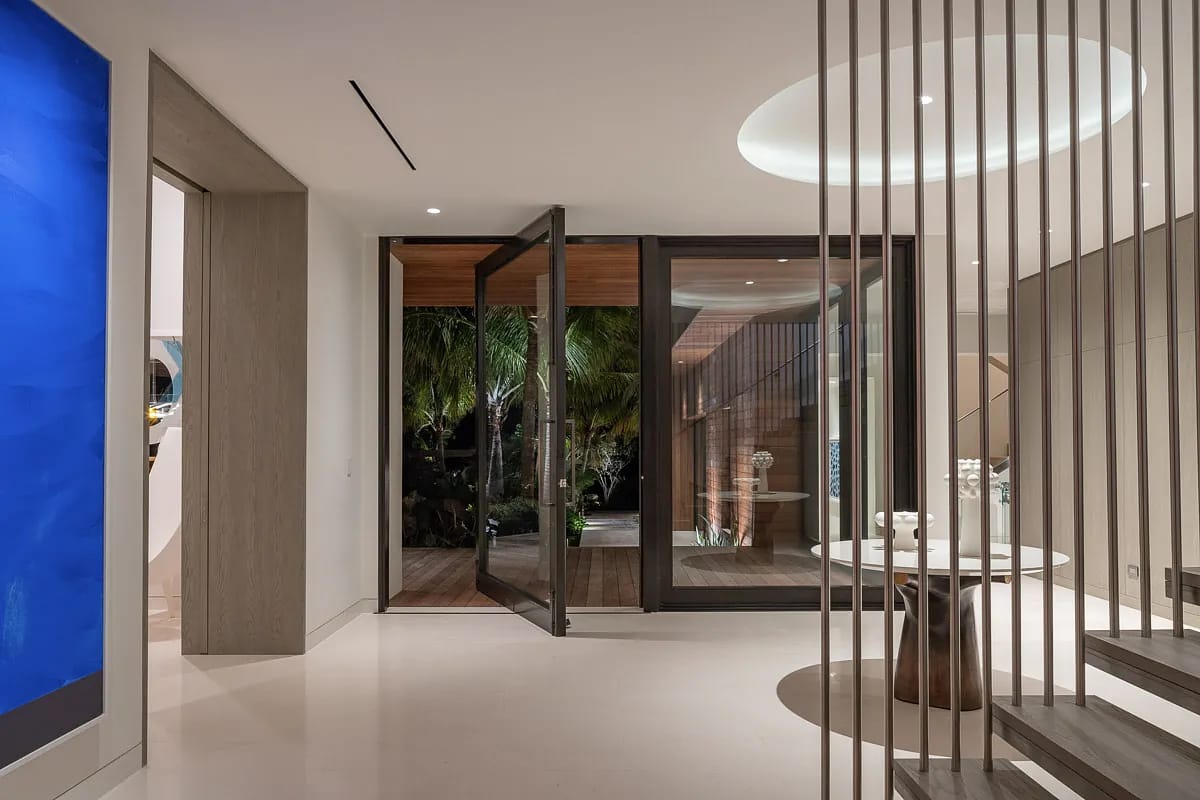 Foyer with glass door, circular table, greenery backdrop, polished floor, modern staircase, and blue artwork.