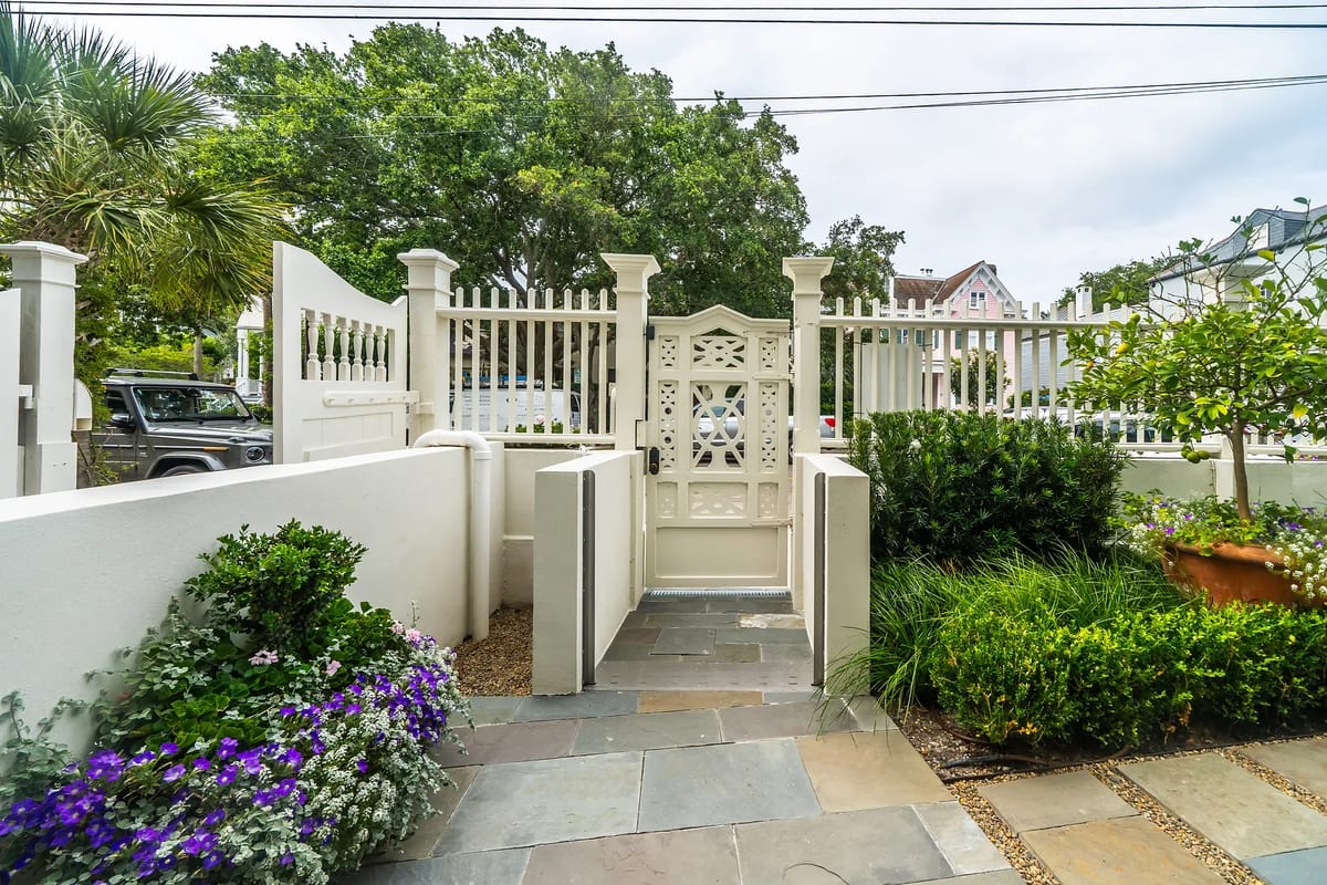 Exterior front area with stone pathway, white wooden gate, and surrounding flowers and plants.