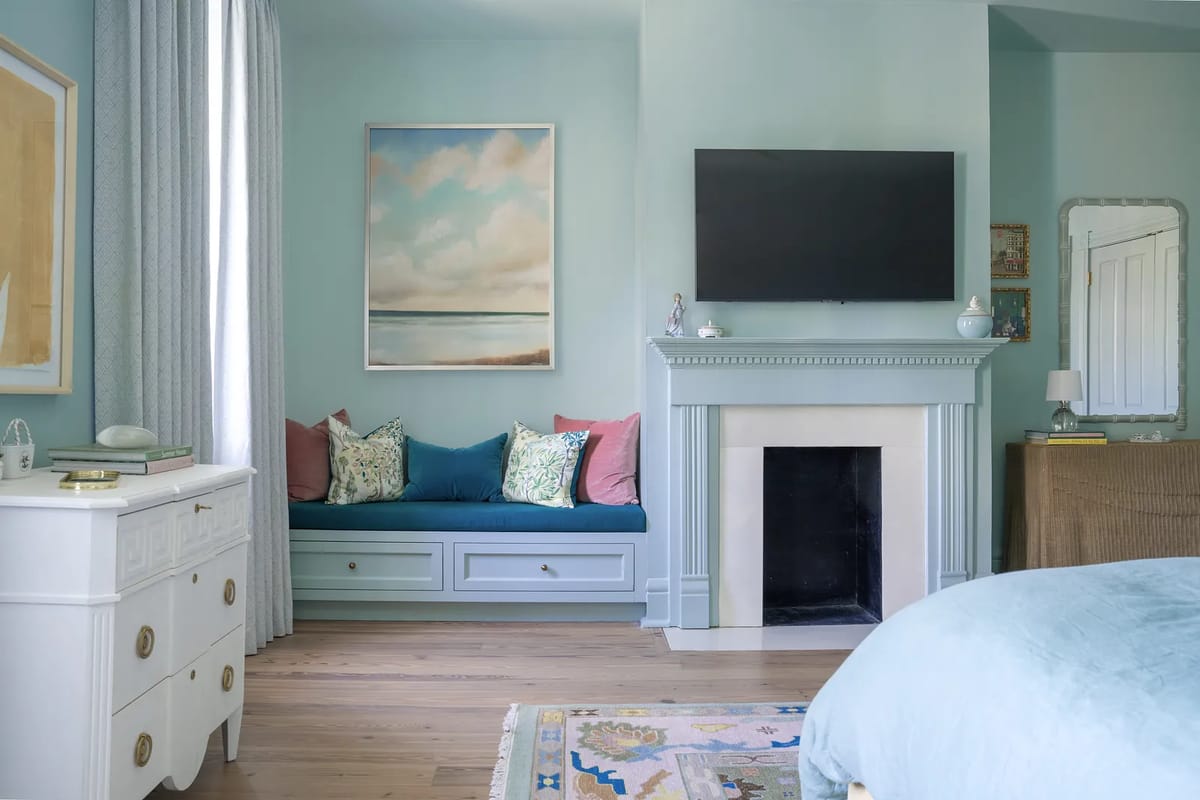 Bedroom with blue walls, window seat, mounted TV, white fireplace, and area rug on wooden floor.