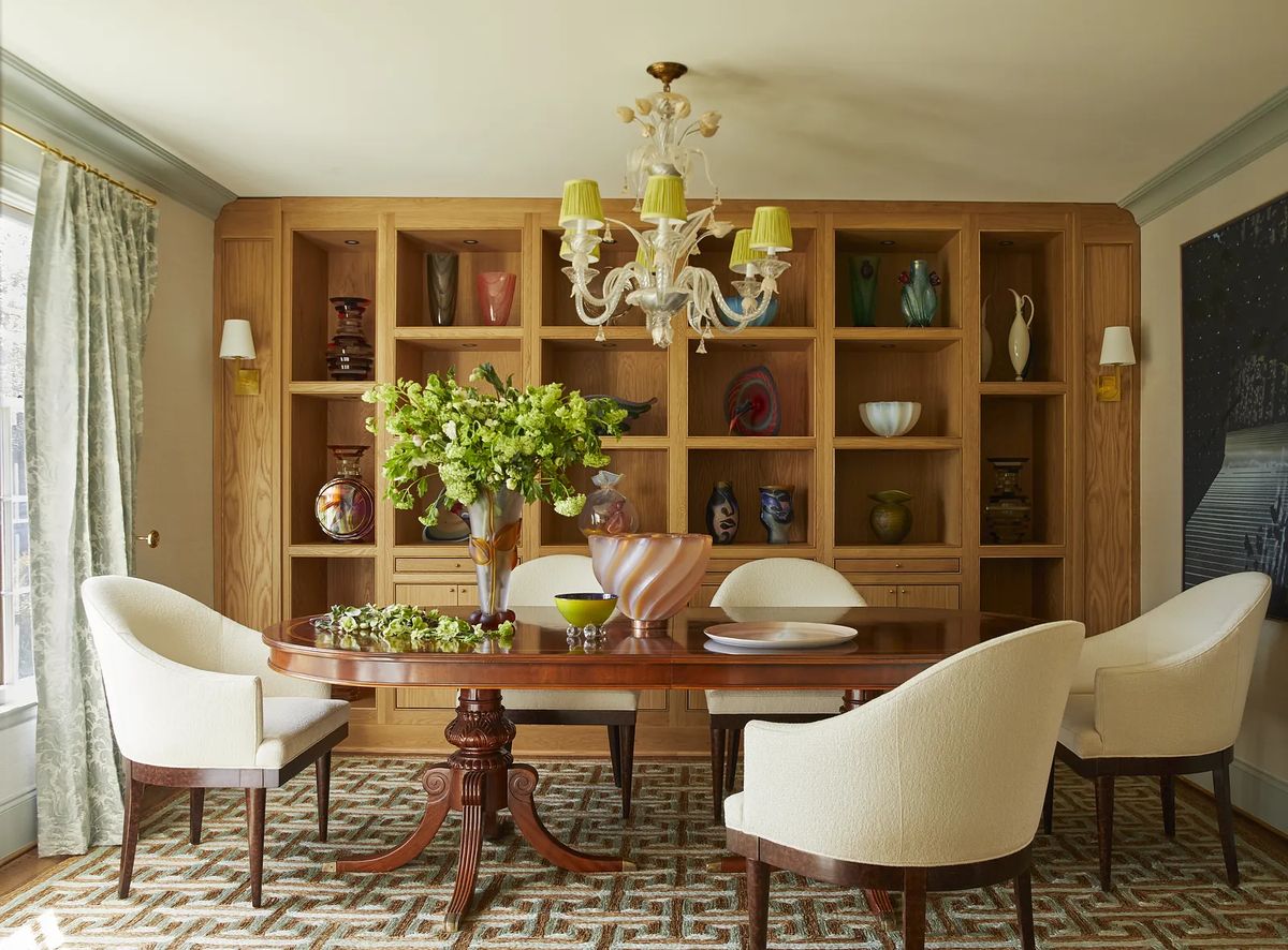 Dining room with round wooden table, white upholstered chairs, shelving unit, and chandelier.