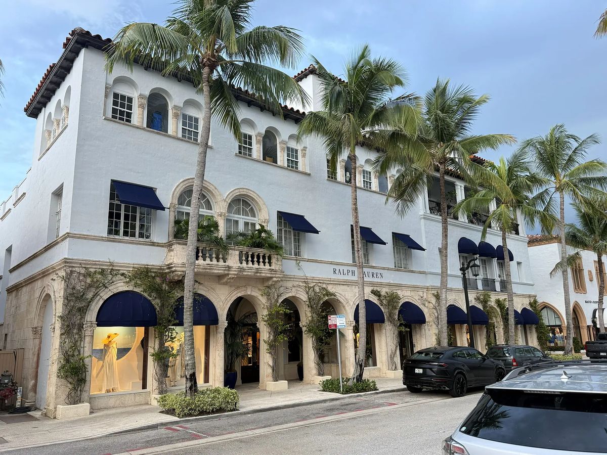 Boutique storefront with white stucco walls, arched windows, black awnings, and palm trees.